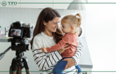 An adult holding a small child stands near a camera on a tripod in a kitchen, with a “TFO Life Academy” logo in the corner.