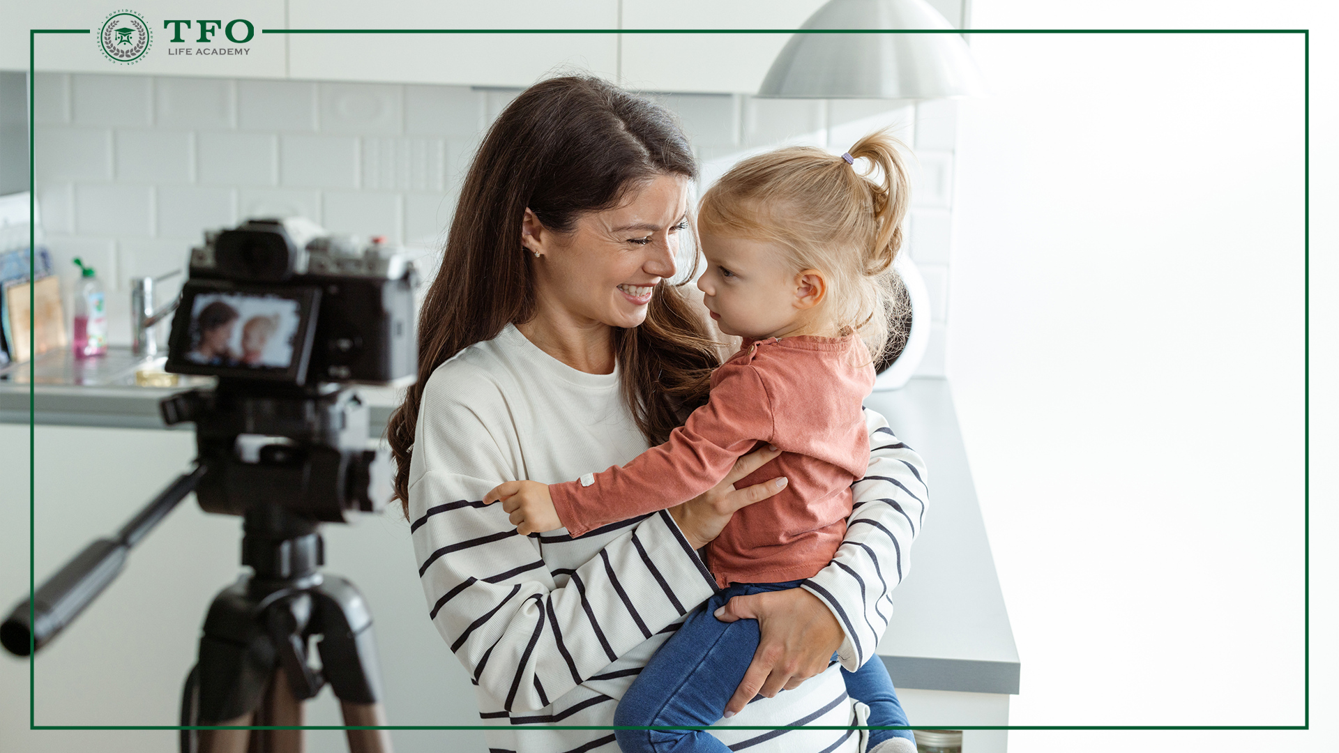 An adult holding a small child stands near a camera on a tripod in a kitchen, with a “TFO Life Academy” logo in the corner.